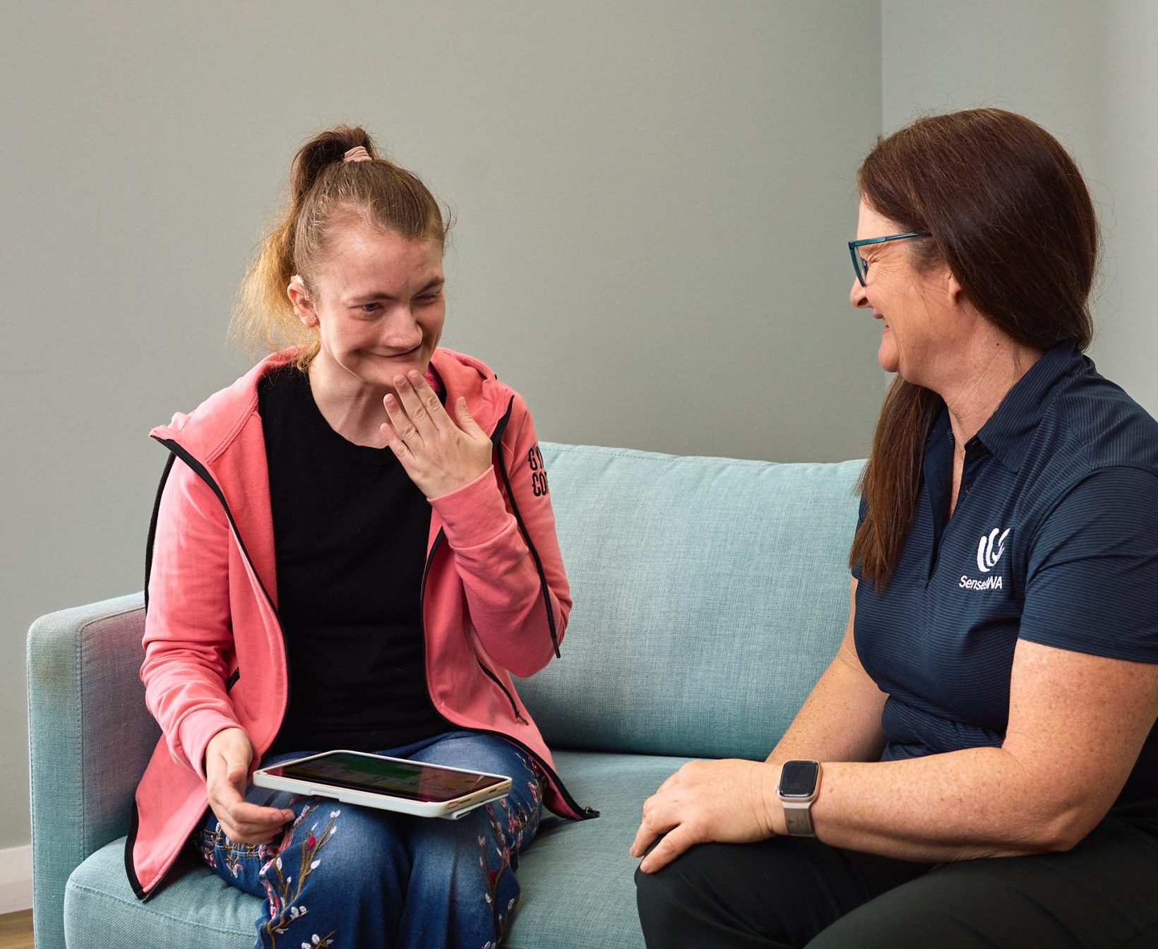 Two women are seated on an aqua couch in a consult room at SensesWA. They are communicating in Key Word Sign.
